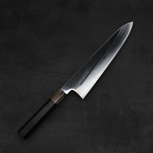 A Satoshi Nakagawa aogami#1 honyaki tall gyuto 240mm knife with a wooden handle and sharp, shiny blade lies on a dark, textured surface.