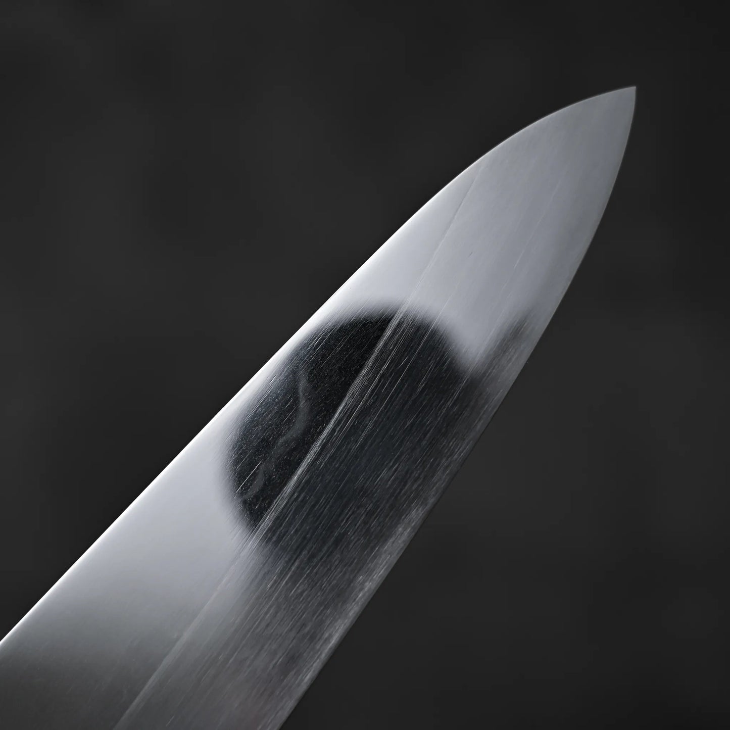 A close-up of the Satoshi Nakagawa aogami#1 honyaki tall gyuto 240mm shows its razor-sharp edge and subtle reflections on the pristine blade, set against a dark, blurred background.
