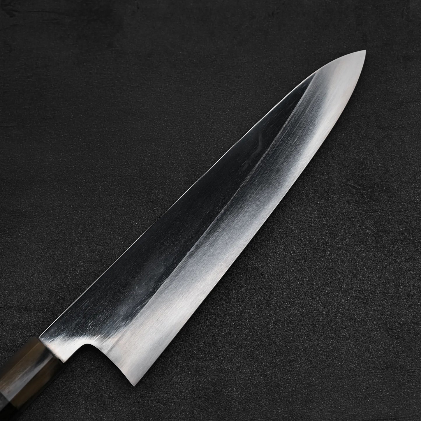 A close-up of the Satoshi Nakagawa aogami#1 honyaki tall gyuto 240mm knife, featuring a sharp blade and dark handle, displayed on a textured black surface.