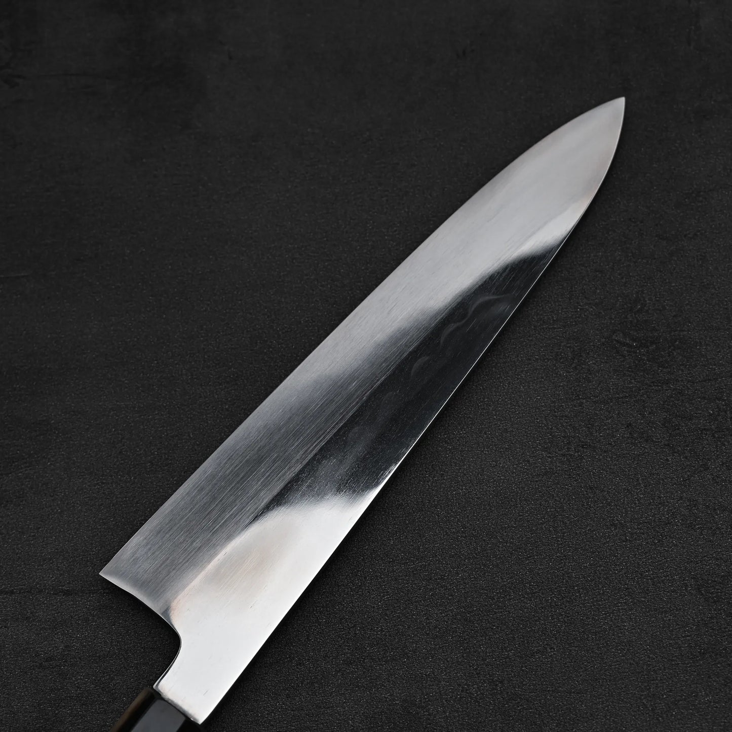 A close-up of the Satoshi Nakagawa aogami#1 honyaki tall gyuto 240mm knife, its shiny blade resting on a dark, textured surface.
