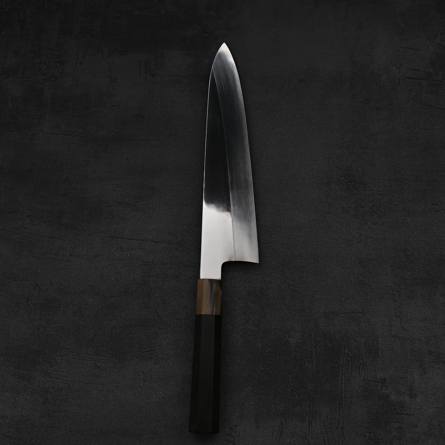 A Satoshi Nakagawa aogami#1 honyaki tall gyuto 240mm Japanese knife with a shiny blade and dark handle rests on a dark textured surface.