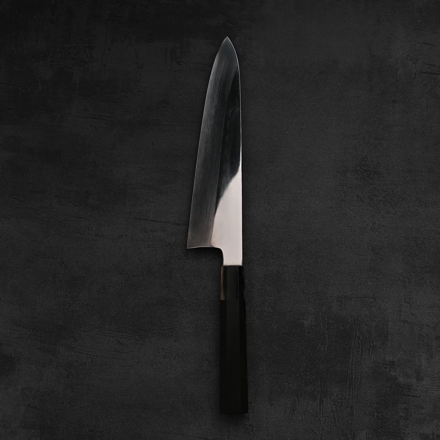 A single Satoshi Nakagawa Nakagawa aogami#1 honyaki tall gyuto 240mm, with a shiny blade and dark handle, stands upright on a textured dark gray surface.