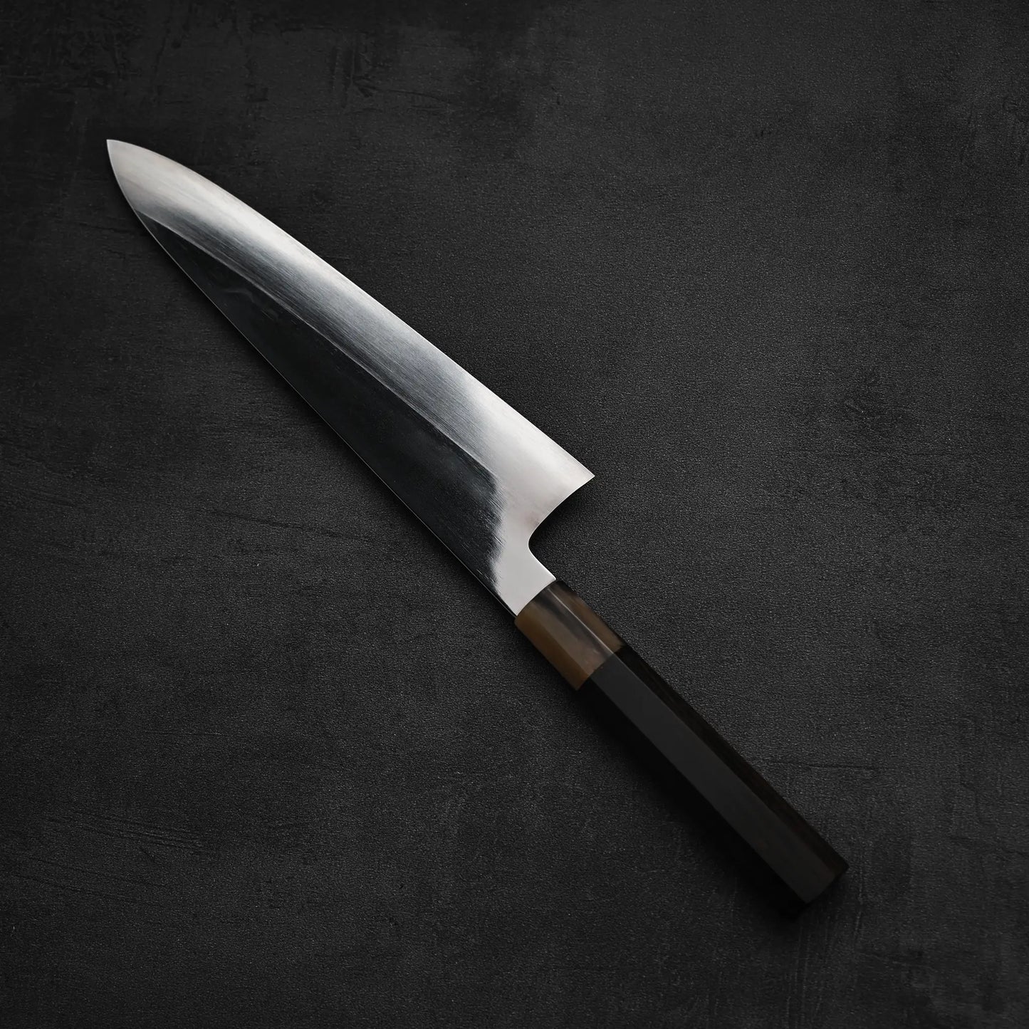 A Satoshi Nakagawa aogami#1 honyaki tall gyuto 240mm knife with a gleaming blade and dark wooden handle rests on a textured black surface.