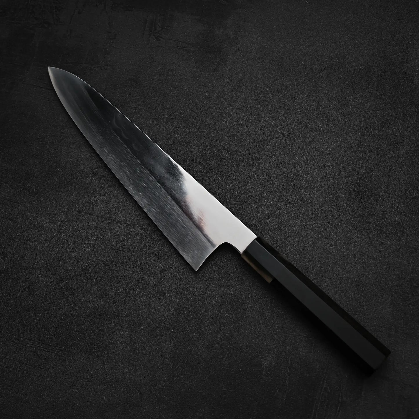 A Satoshi Nakagawa aogami#1 honyaki tall gyuto 240mm with a reflective stainless steel blade and black handle is displayed on a dark, textured surface.