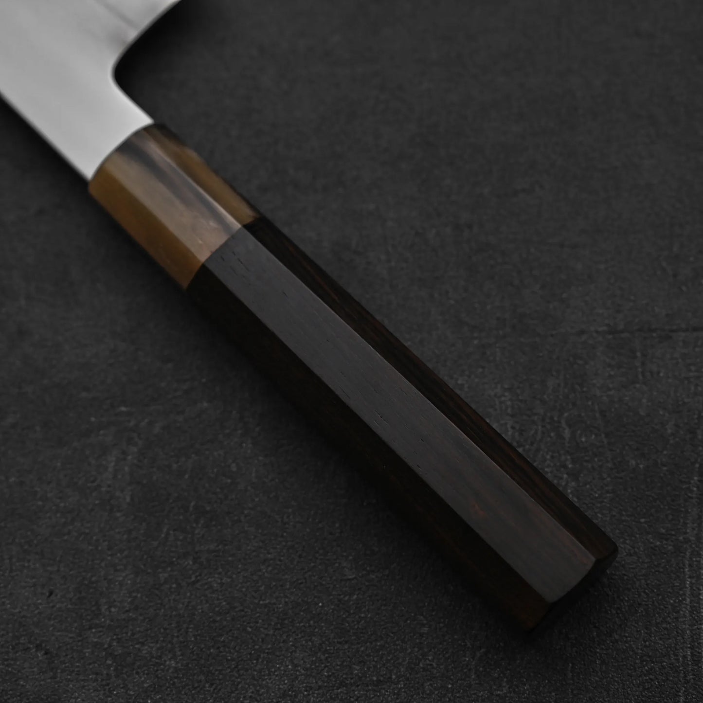 Close-up of a Japanese knife handle crafted from dark and light wood, resting on a textured surface. The blade of this Satoshi Nakagawa aogami#1 honyaki tall gyuto 240mm is partially visible in the upper left corner.