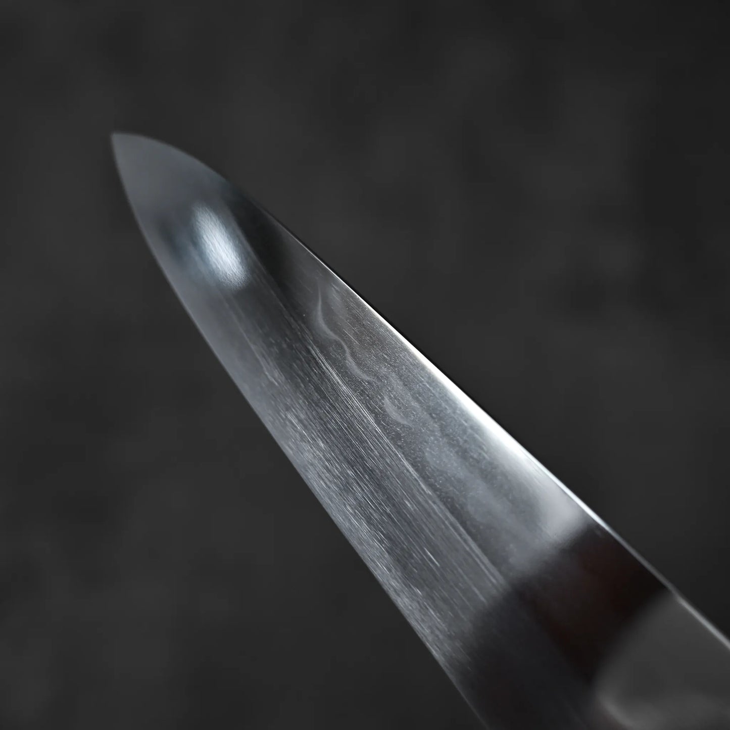 Close-up of the Satoshi Nakagawa aogami#1 honyaki tall gyuto 240mm blade, featuring a smooth, reflective surface with subtle texture and a sharp edge set against a dark, blurred background.