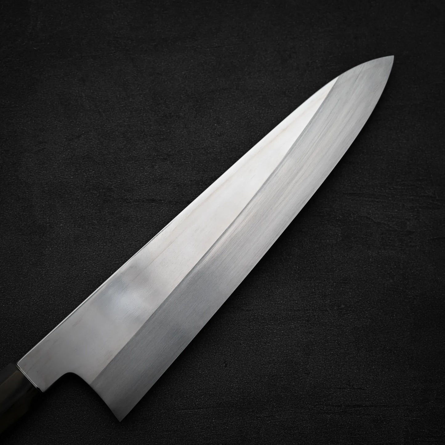 A close-up of the Satoshi Nakagawa Nakagawa aogami#1 honyaki tall gyuto 240mm chef’s knife, featuring a shiny stainless steel blade, positioned diagonally on a dark, textured background.