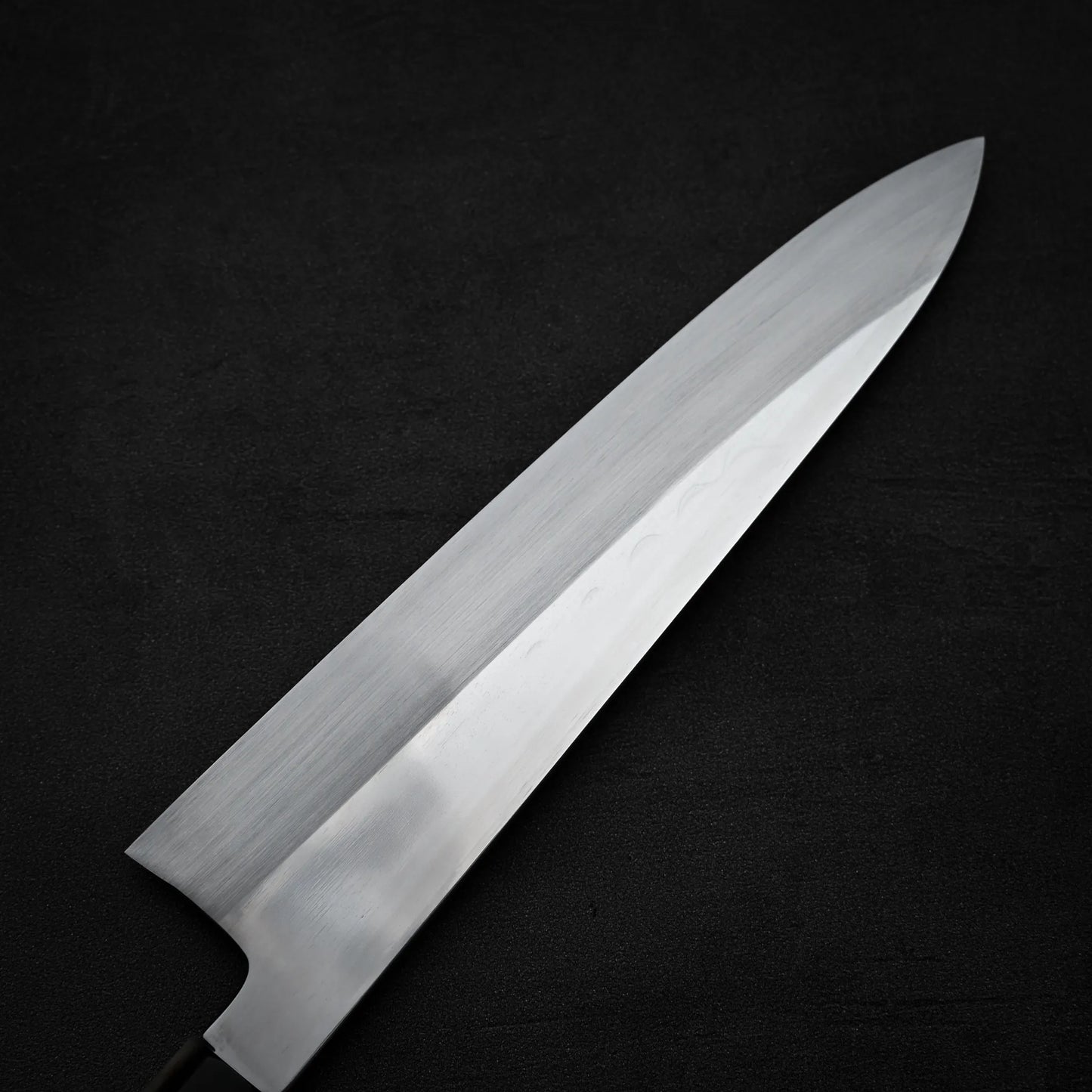 A close-up of a Satoshi Nakagawa Nakagawa aogami#1 honyaki tall gyuto 240mm, featuring a sharp silver blade, positioned diagonally on a dark, textured surface.