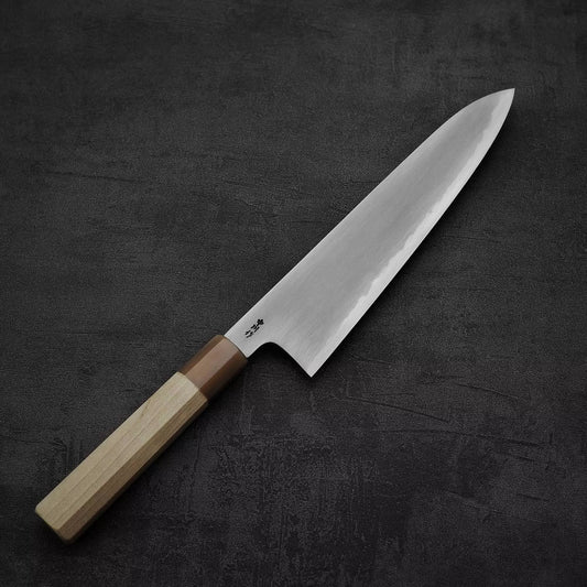 A Satoshi Nakagawa bokashi shirogami#1 tall gyuto 240mm chef knife with a wooden handle and gleaming blade rests elegantly on a dark textured surface.