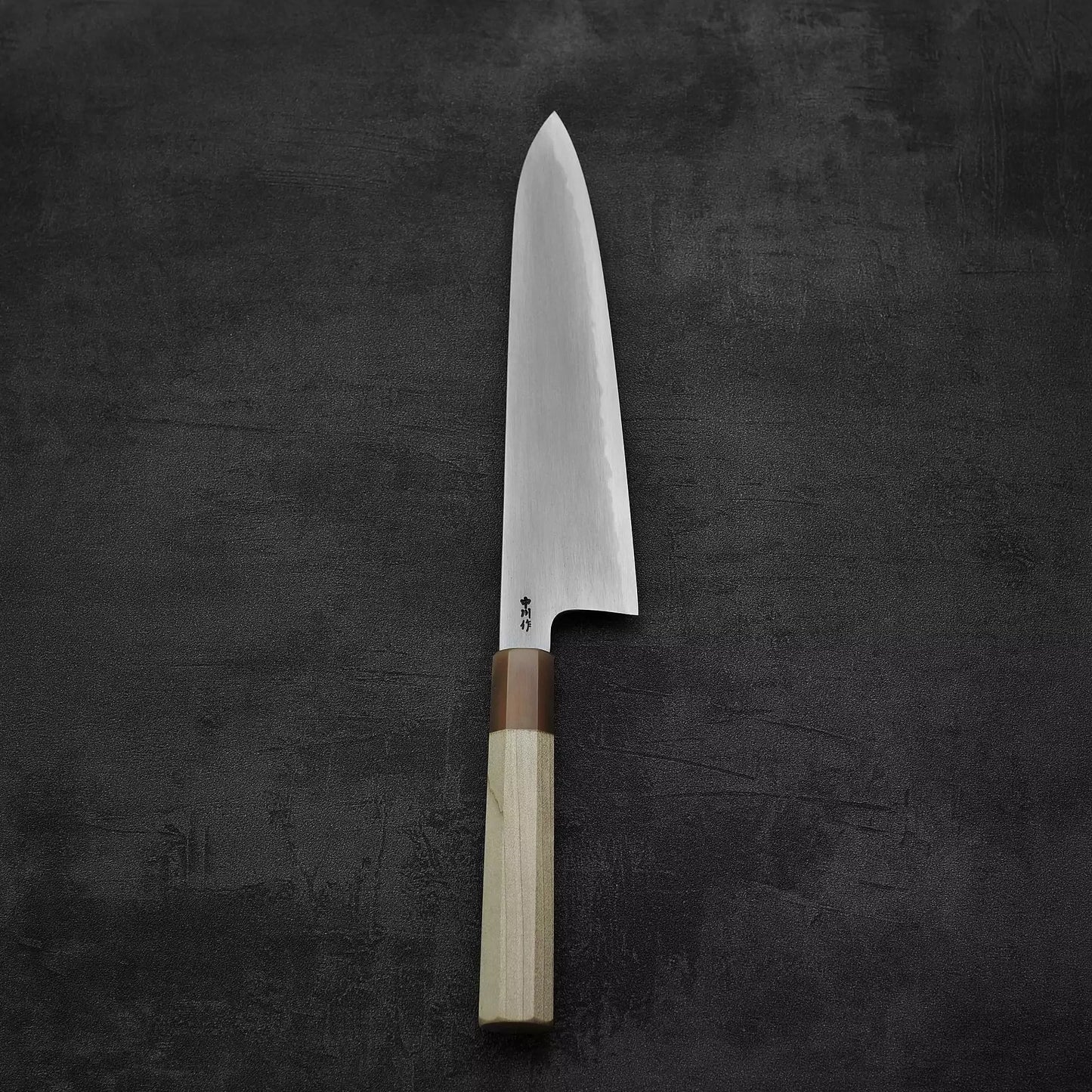 The Satoshi Nakagawa bokashi shirogami#1 tall gyuto 240mm, featuring a gleaming blade and wooden handle, rests elegantly on a dark, textured surface.