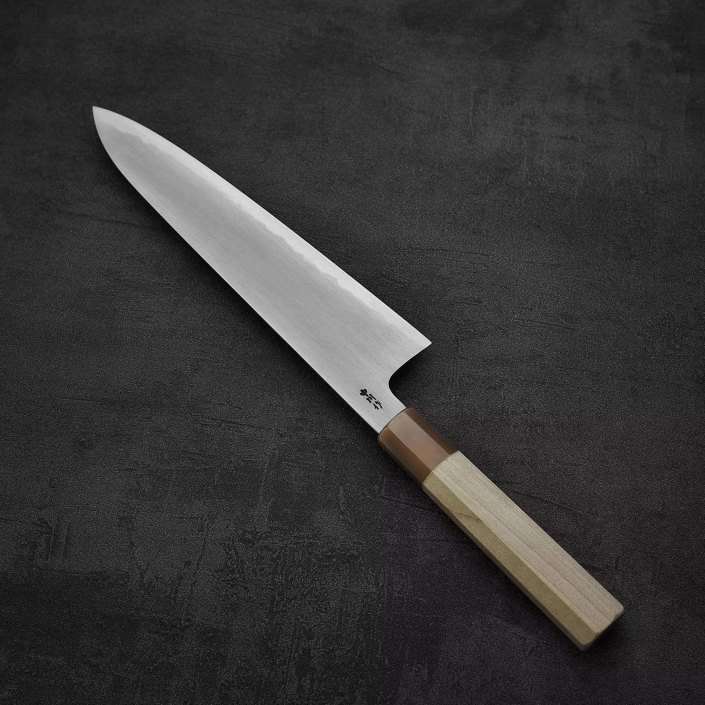 A Satoshi Nakagawa bokashi shirogami#1 tall gyuto 240mm with a shiny pointed blade and wooden handle rests on a dark, textured surface.