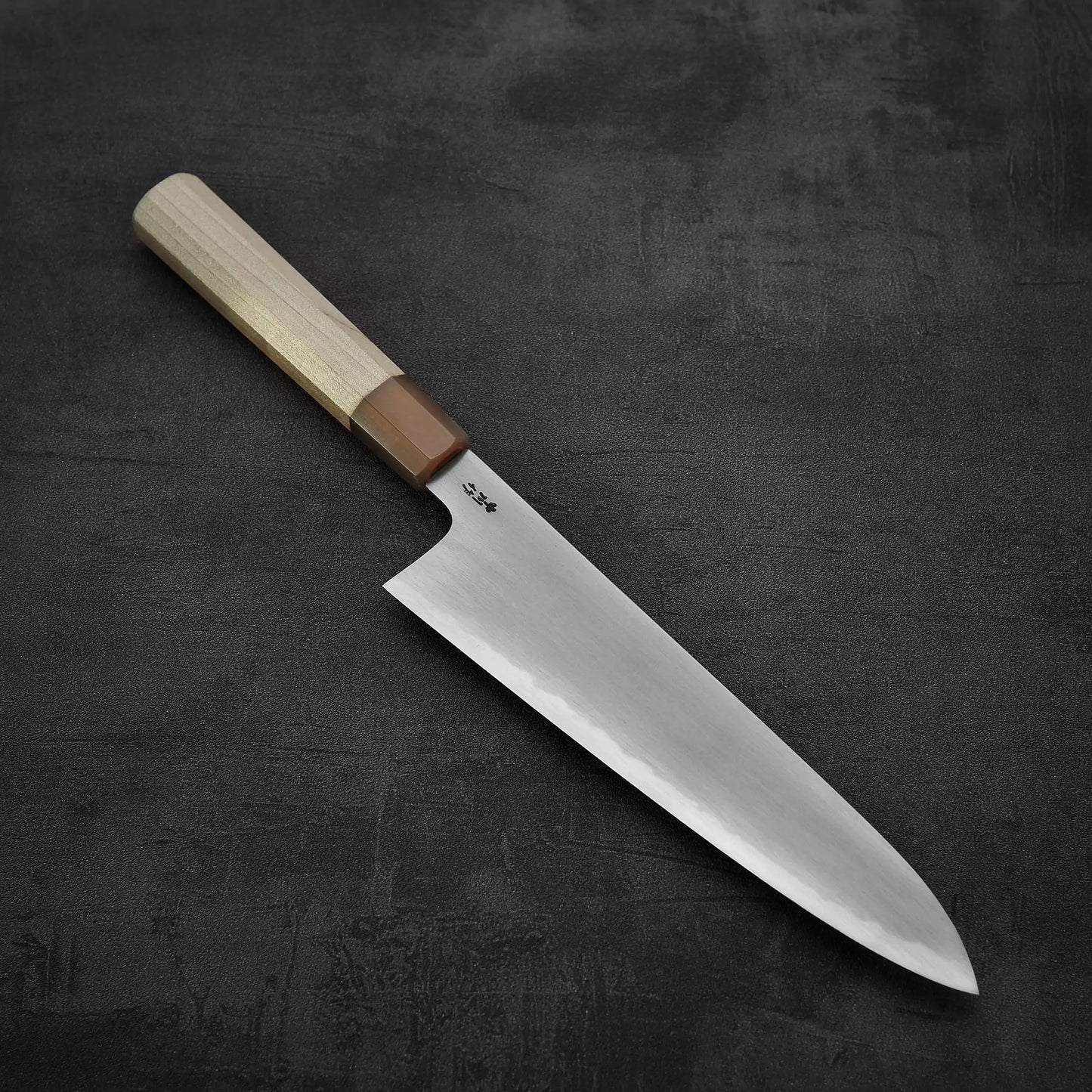 A Satoshi Nakagawa bokashi shirogami#1 tall gyuto 240mm with a wooden handle lies diagonally on a dark textured surface, its polished blade pointing to the lower right.