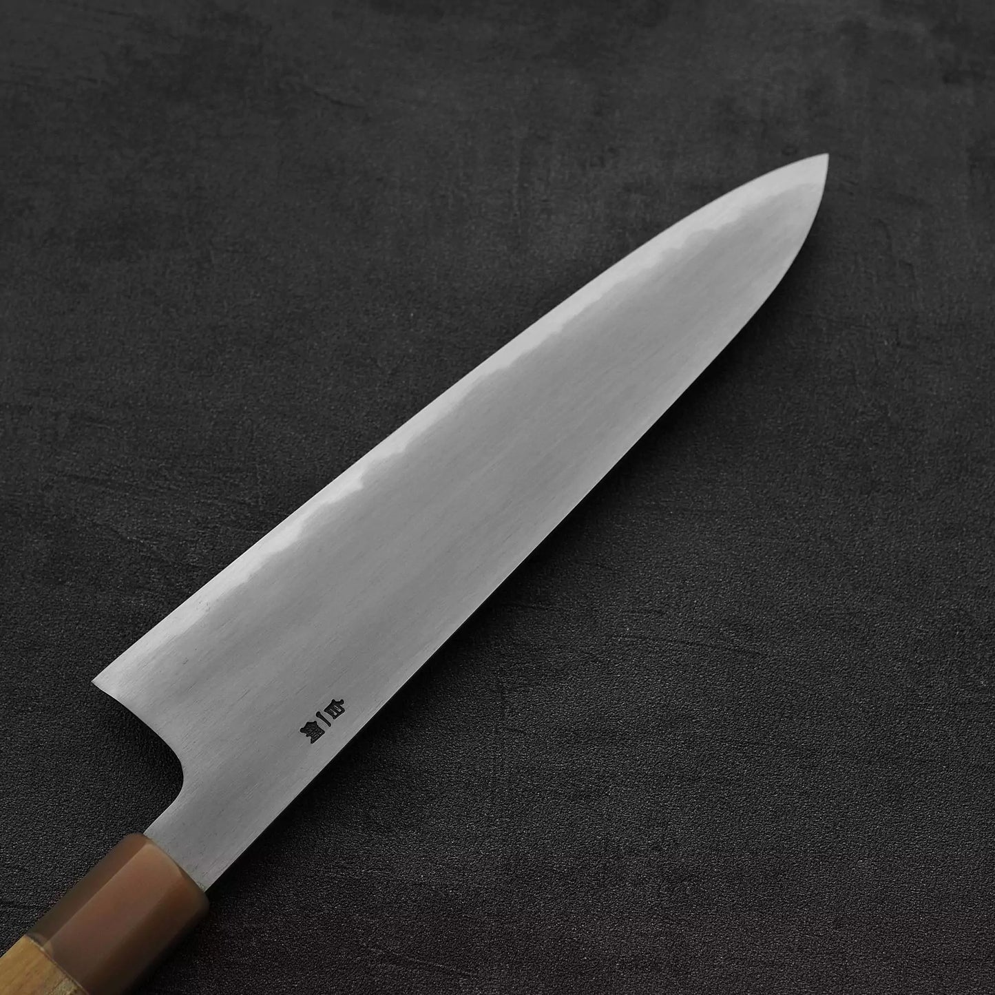 A close-up of the Satoshi Nakagawa bokashi shirogami#1 tall gyuto 240mm features its wooden handle and shiny, sharp blade, resting on a dark textured surface.