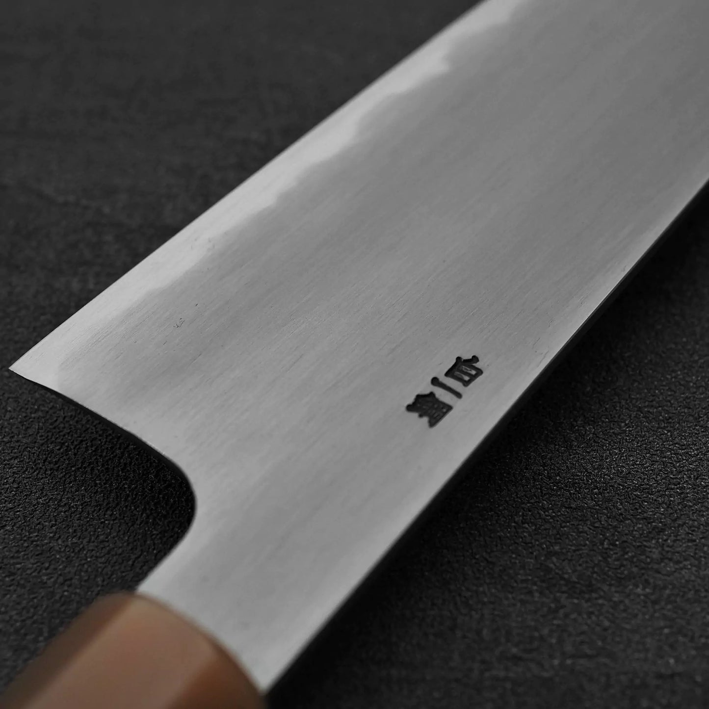 Close-up of the Satoshi Nakagawa bokashi shirogami#1 tall gyuto 240mm, showing its wooden handle and engraved shirogami#1 steel blade with a subtle bokashi finish, resting on a dark textured surface.