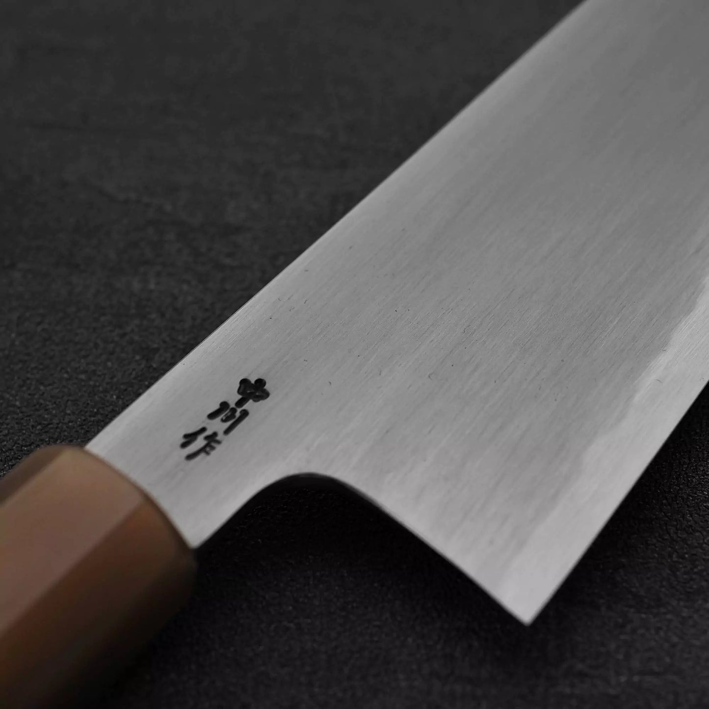 A close-up of the Satoshi Nakagawa bokashi shirogami#1 tall gyuto 240mm, a Japanese kitchen knife with a wooden handle and engraved blade, set against a dark textured background.