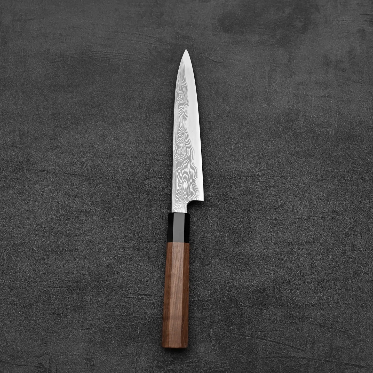 The Hatsukokoro Nakagawa Damascus Aogami#1 Petty Knife 150mm, featuring a stunning Damascus finish and wooden handle, rests on a dark textured surface, showcasing its refined craftsmanship.