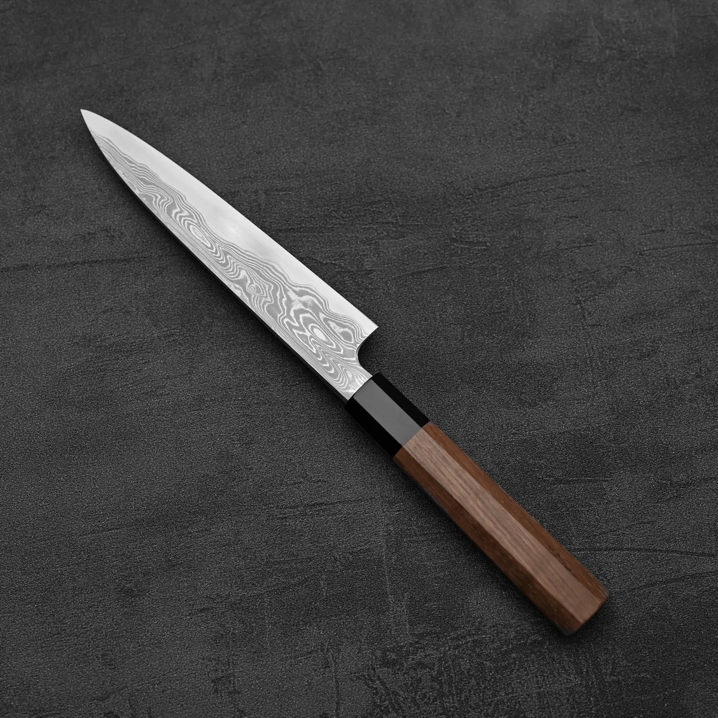 The Hatsukokoro Nakagawa Damascus Aogami#1 Petty Knife 150mm, featuring a wooden handle and Damascus-finish blade, rests on a dark textured surface.