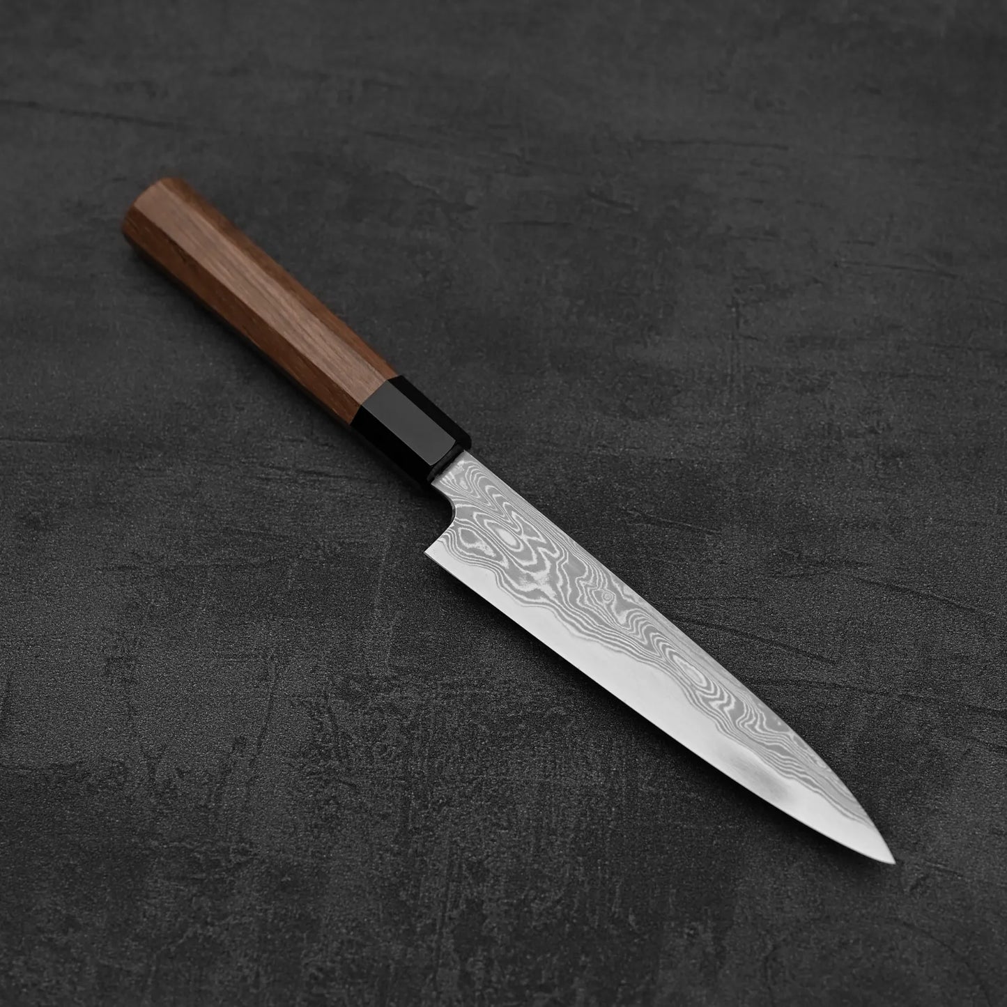 The Hatsukokoro Nakagawa Damascus Aogami#1 Petty Knife 150mm, featuring a wooden handle, rests on a dark textured surface, highlighting its beautifully patterned Damascus blade.