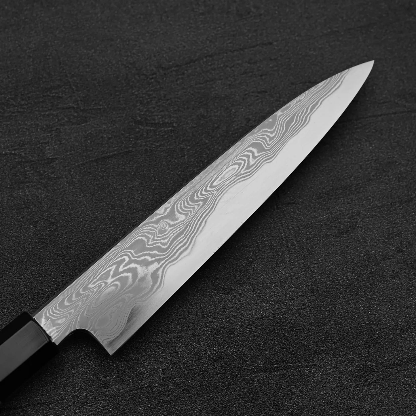The Hatsukokoro Nakagawa damascus aogami#1 petty knife 150mm features a striking Damascus finish and black handle, resting on a dark textured surface with its blade’s fine patterns showcasing elegance and precision.
