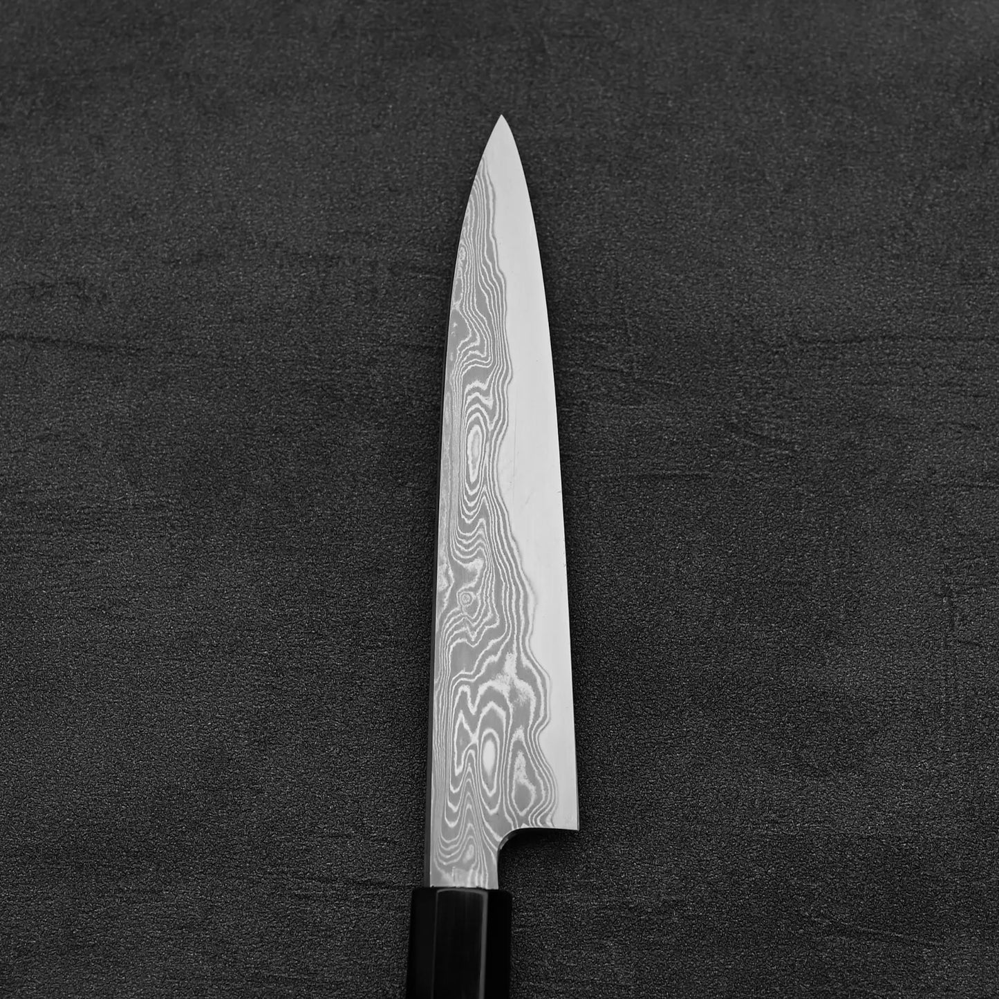 A close-up of the Hatsukokoro Nakagawa damascus aogami#1 petty knife 150mm, featuring wavy Damascus patterns on the blade, placed on a dark textured surface.