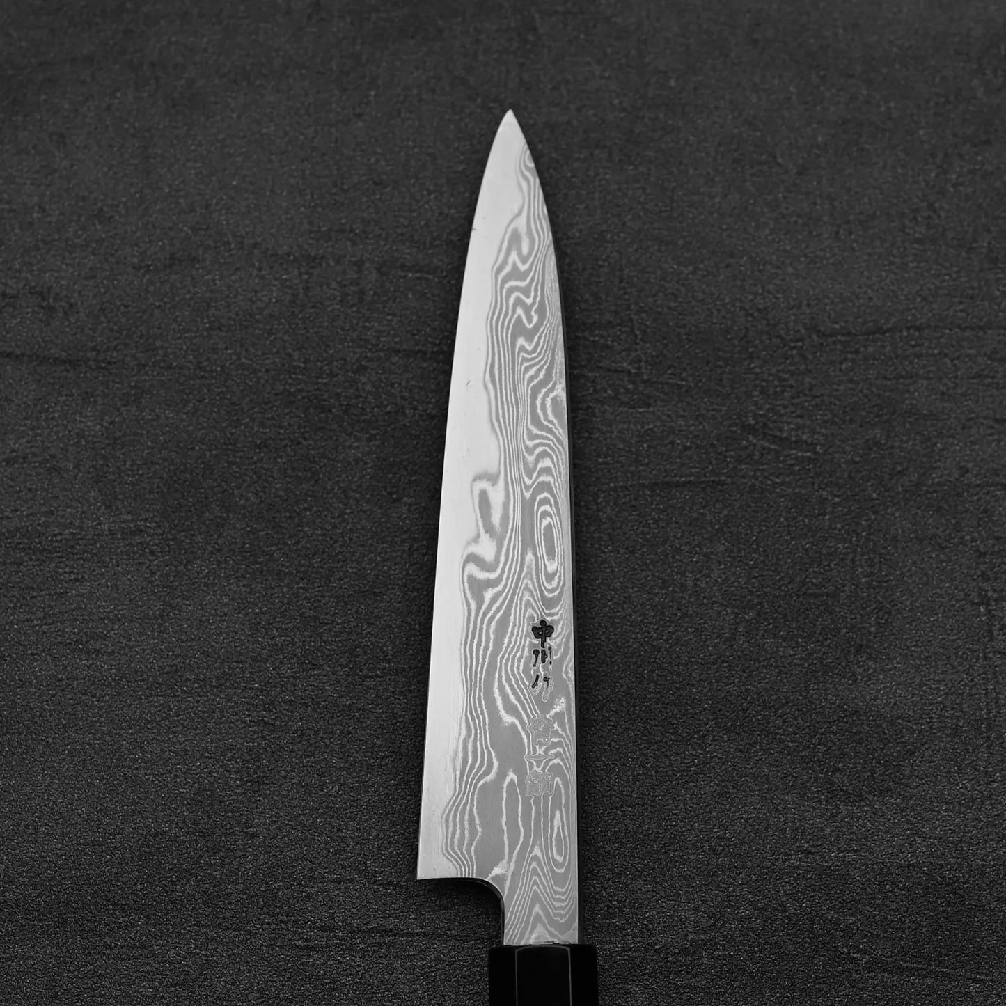 The Hatsukokoro Nakagawa damascus aogami#1 petty knife 150mm, featuring distinctive wavy Damascus patterns on the blade, is photographed on a dark, textured surface with the handle partially visible at the bottom.