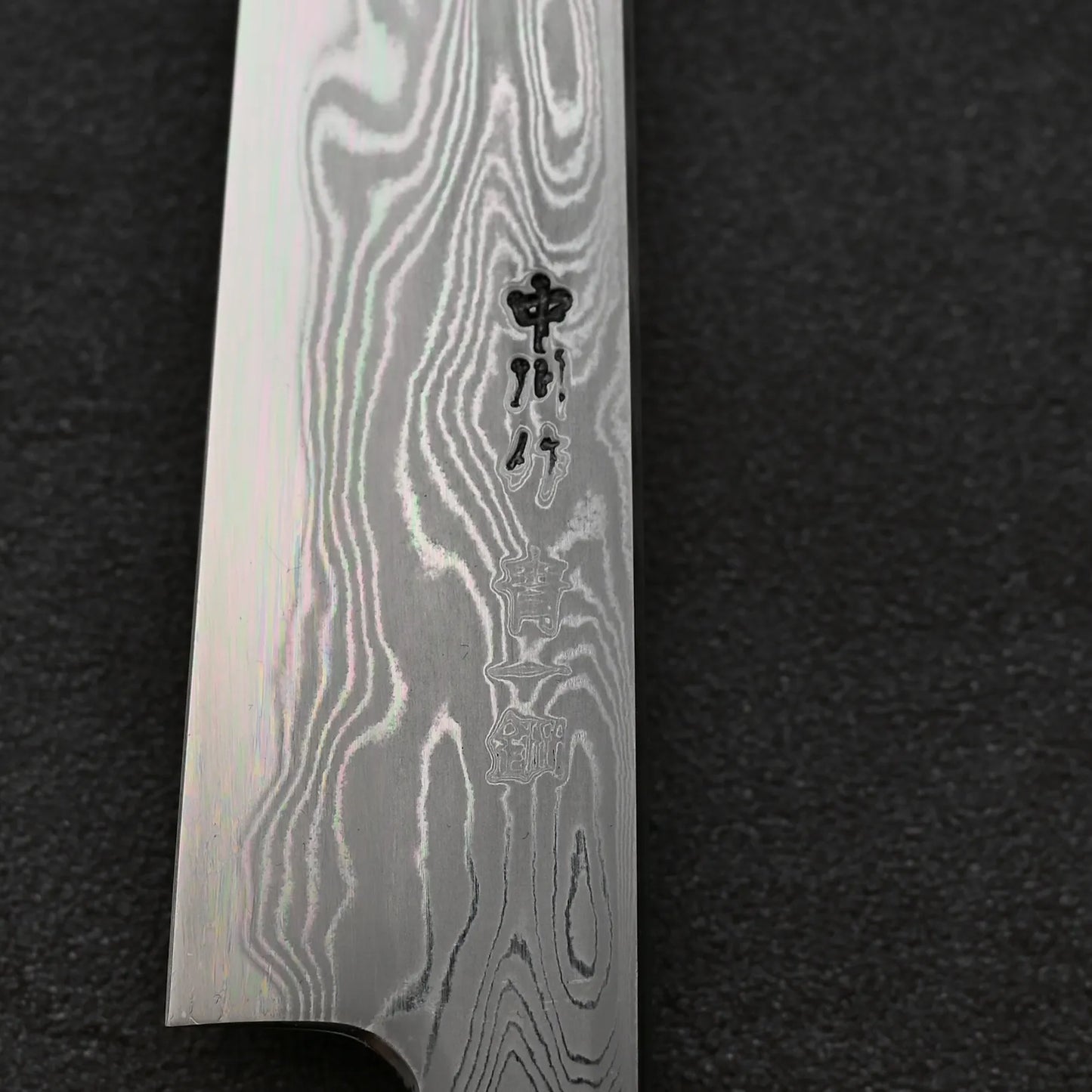 Close-up of the Hatsukokoro Nakagawa damascus aogami#1 petty knife 150mm blade showcasing its striking Damascus finish and engraved Japanese characters, set against a dark textured background.