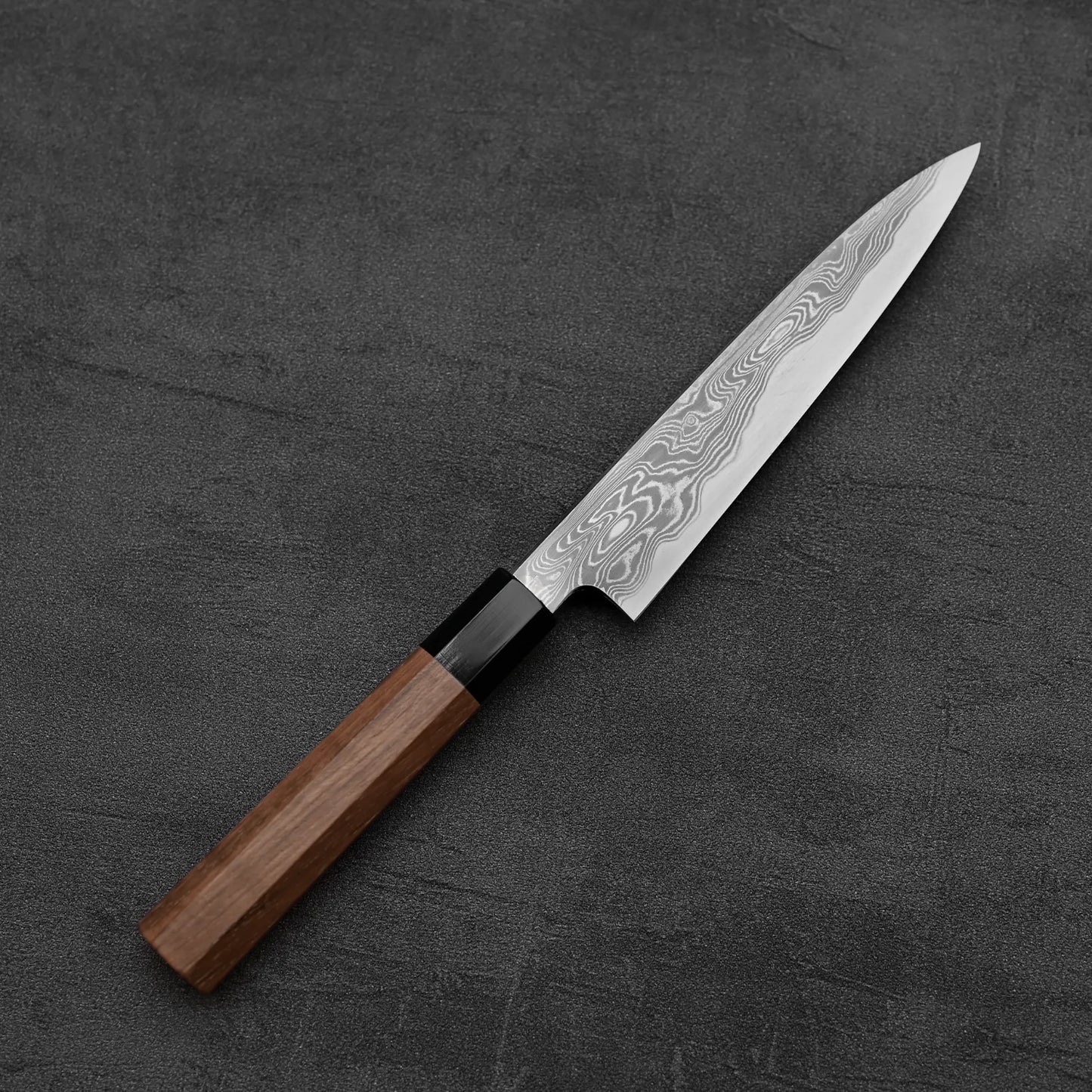 The Hatsukokoro Nakagawa Damascus Aogami#1 Petty Knife 150mm features a wooden handle and wavy-patterned Damascus blade, showcased on a dark textured surface.
