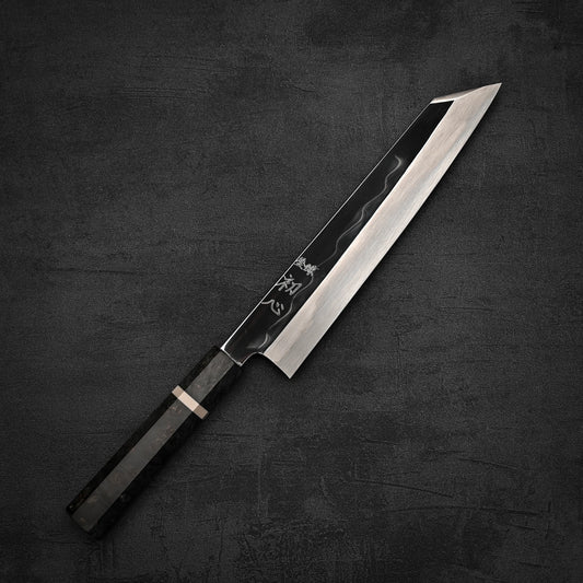 A Hatsukokoro Nakagawa shirogami#2 mizuhonyaki kiritsuke gyuto 240mm chef’s knife with a sharp, angled blade and dark patterned handle rests on a textured dark gray surface.
