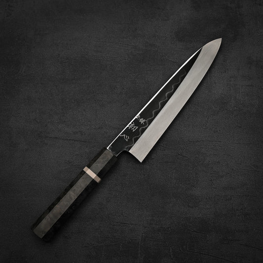 The Hatsukokoro Nakagawa shirogami#3 honyaki gyuto 210mm, with a black patterned blade and dark handle, lies diagonally on a textured dark gray surface.