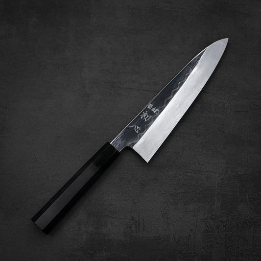 The Satoshi Nakagawa shirogami#3 honyaki tall gyuto 210mm features a black handle and a shiny, hammered steel blade, displayed on a dark textured surface.