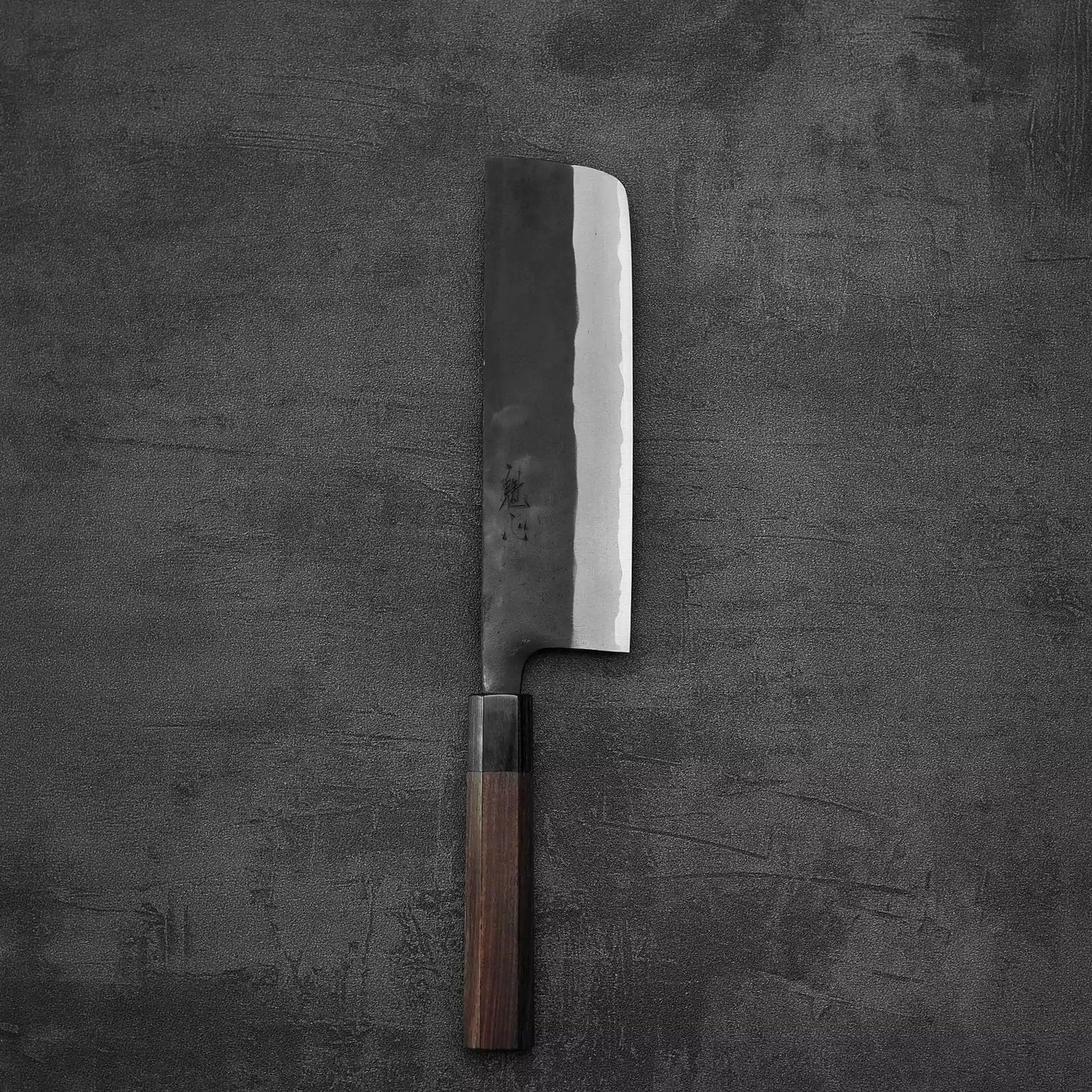 A Nao Yamamoto kurouchi aogami#2 nakiri 180mm knife with excellent edge retention and a dark wooden handle rests on a textured dark gray surface.