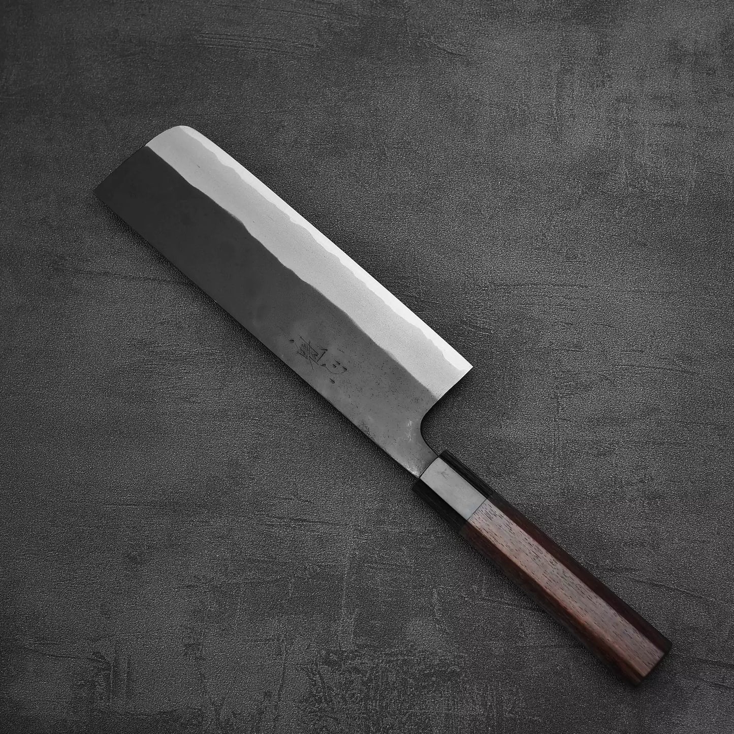 The Nao Yamamoto kurouchi aogami#2 nakiri 180mm, a Japanese vegetable knife by Nao Yamamoto, features a rectangular blade and dark wooden handle resting on a textured dark gray surface.