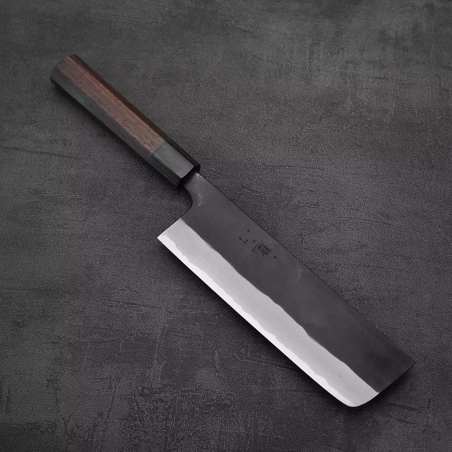 The Nao Yamamoto kurouchi aogami#2 nakiri 180mm, known for its rectangular blade and dark wooden handle, showcases exceptional edge retention as it rests on a textured dark grey surface.