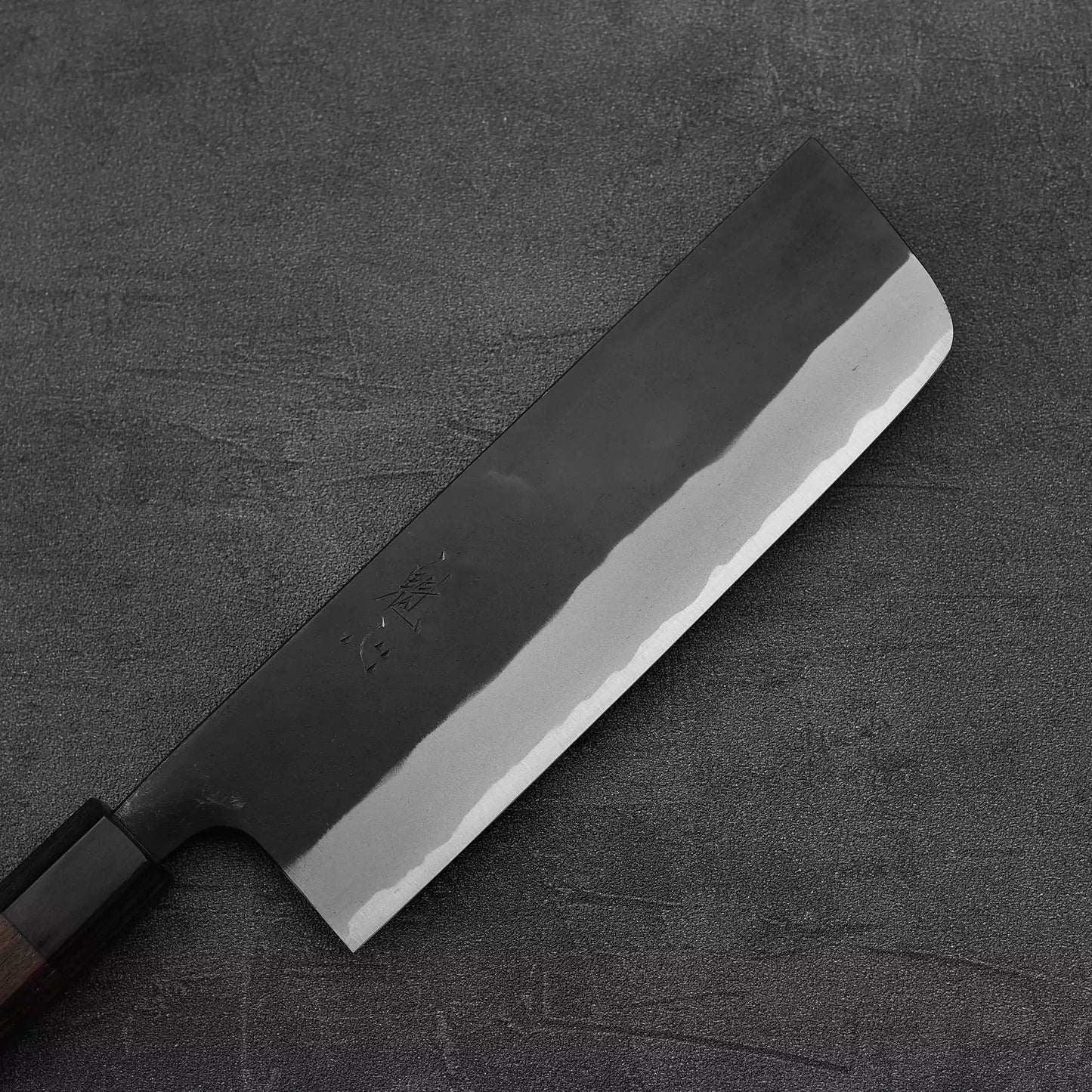 The Nao Yamamoto kurouchi aogami#2 nakiri 180mm, a Japanese kitchen knife with a straight edge and rectangular blade, is placed on a dark textured surface—renowned for superior edge retention.