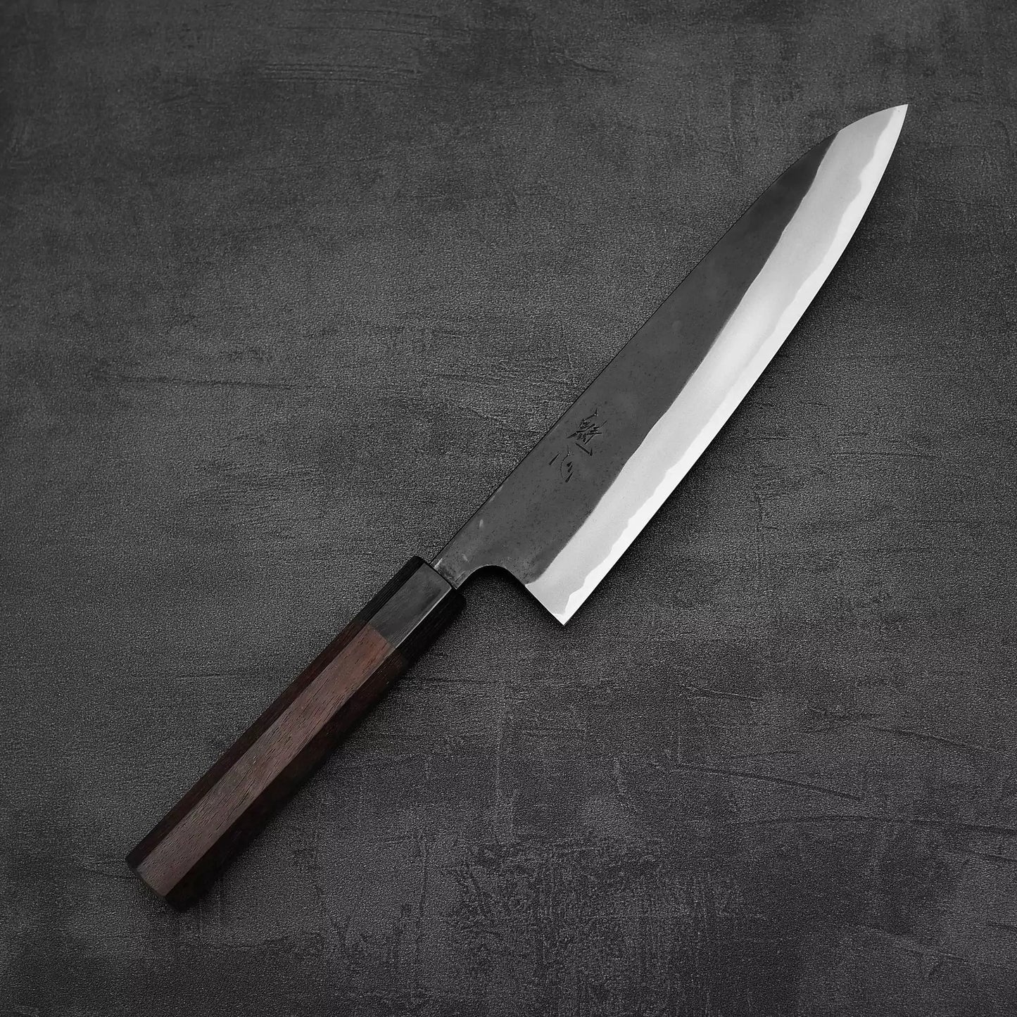A Nao Yamamoto kurouchi shirogami#2 gyuto 210mm with a rosewood handle and wide, polished blade rests diagonally on a dark textured surface.