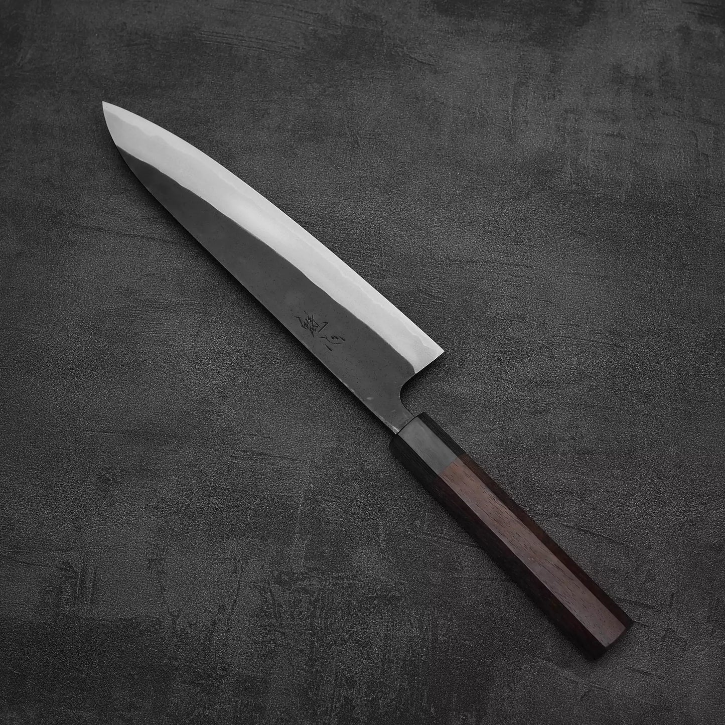 A Nao Yamamoto kurouchi shirogami#2 gyuto 210mm with a polished blade and dark rosewood handle rests on a textured dark gray surface.