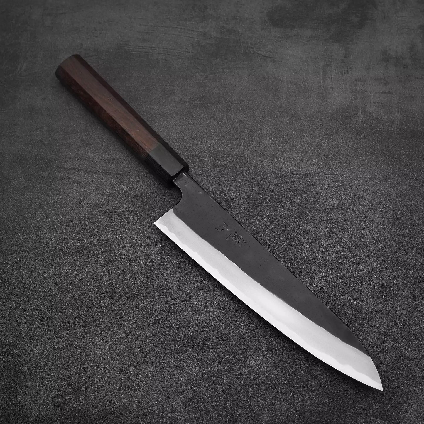 A Nao Yamamoto kurouchi shirogami#2 gyuto 210mm chef’s knife with a rosewood handle rests on a dark, textured surface.