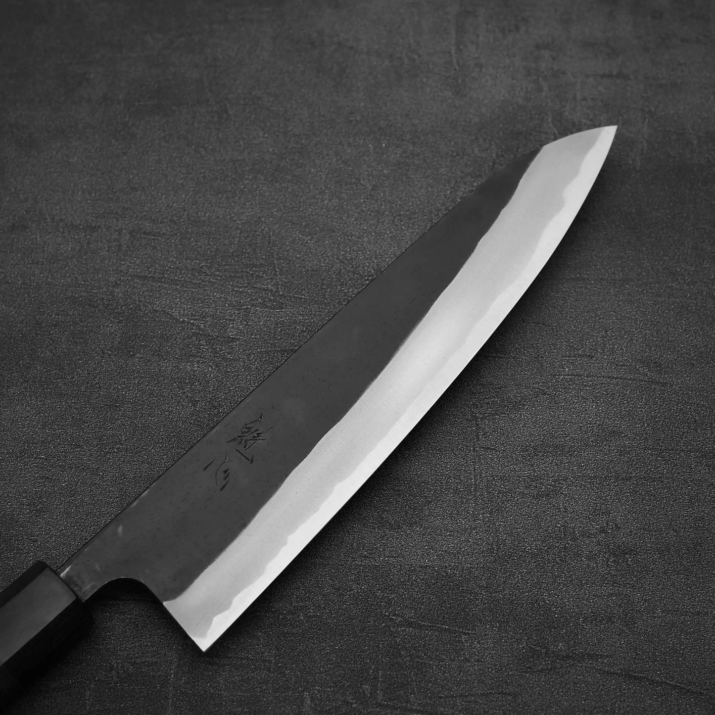 The Nao Yamamoto kurouchi shirogami#2 gyuto 210mm, with a sharp blade and black rosewood handle, rests on a dark textured surface.