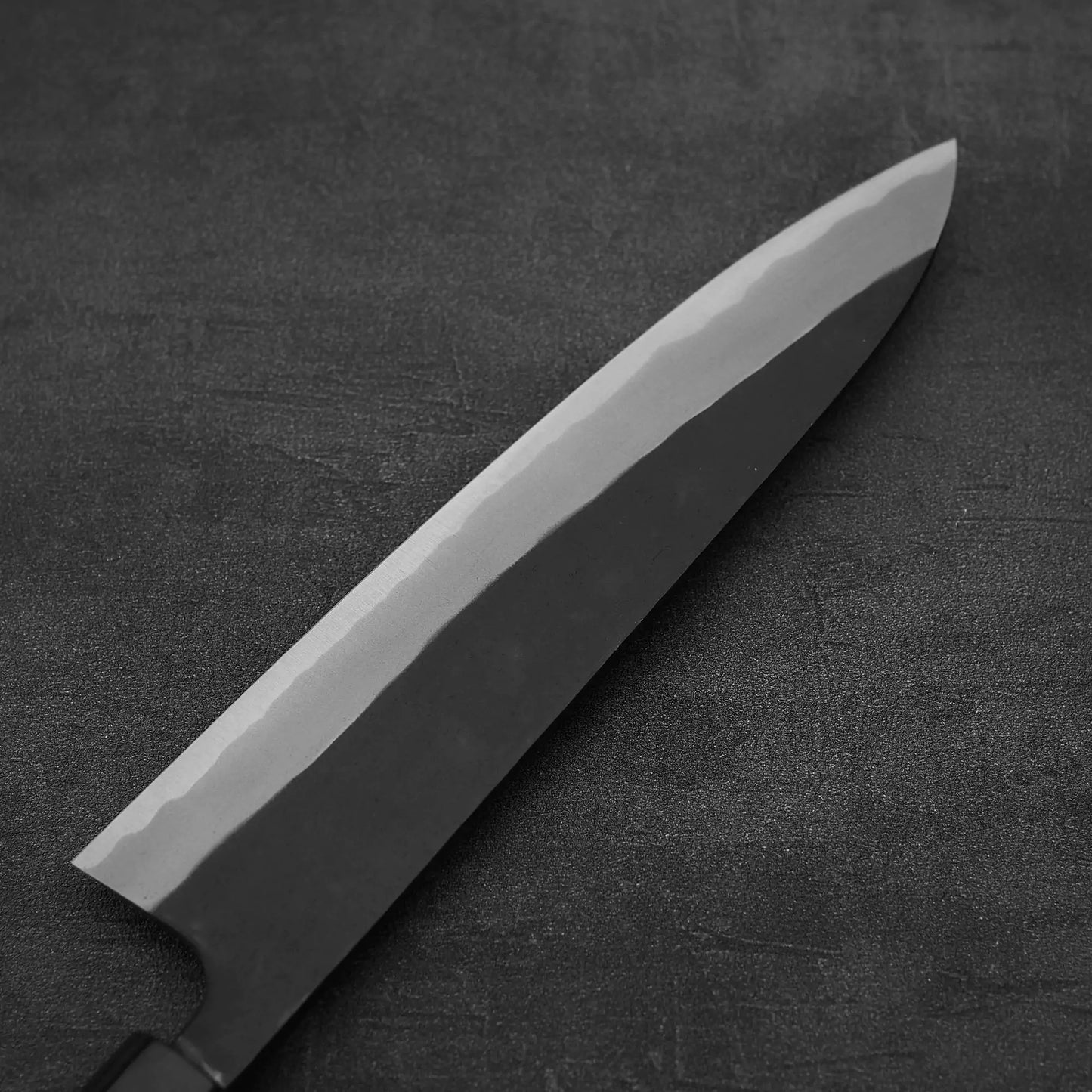 A close-up of the Nao Yamamoto kurouchi shirogami#2 gyuto 210mm, showcasing its dark blade, sharp edge, and smooth rosewood handle, resting on a textured dark surface.