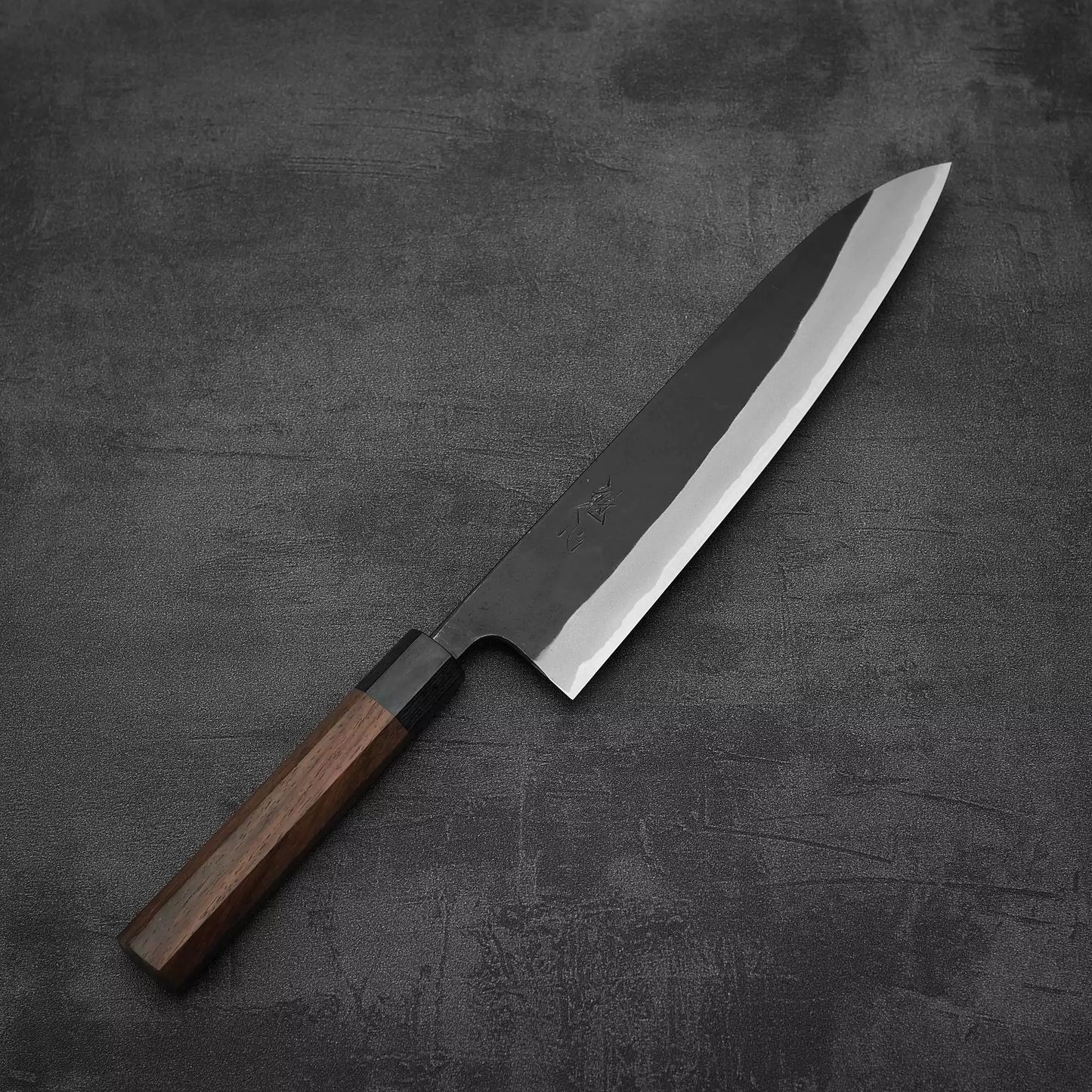 The Nao Yamamoto kurouchi shirogami#2 gyuto 240mm features a dark wooden handle and a sharp, shiny blade, elegantly displayed on a dark textured surface.