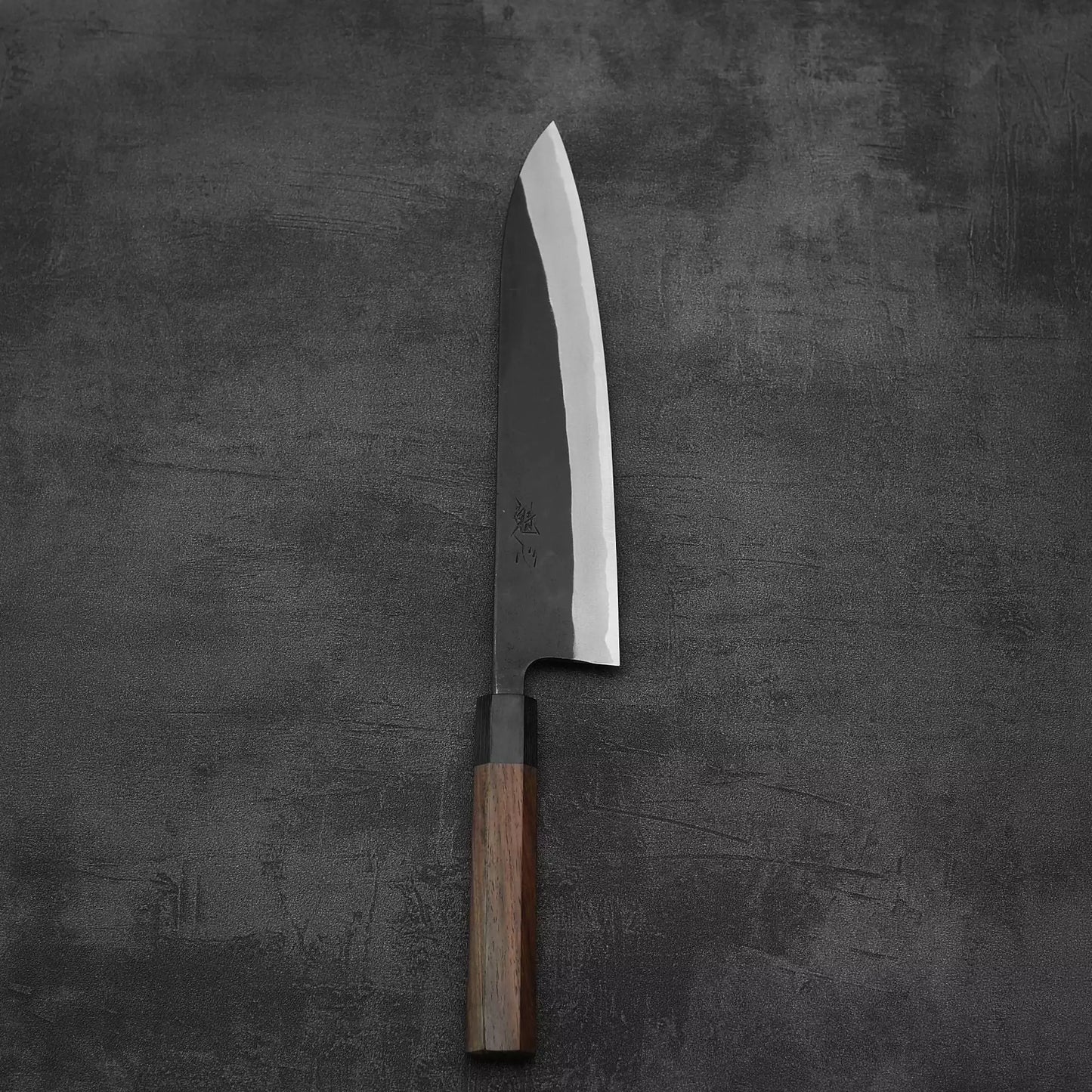 The Nao Yamamoto kurouchi shirogami#2 gyuto 240mm, a handmade Japanese knife with a sharp, wide shirogami#2 steel blade and wooden handle, rests on a dark, textured surface.