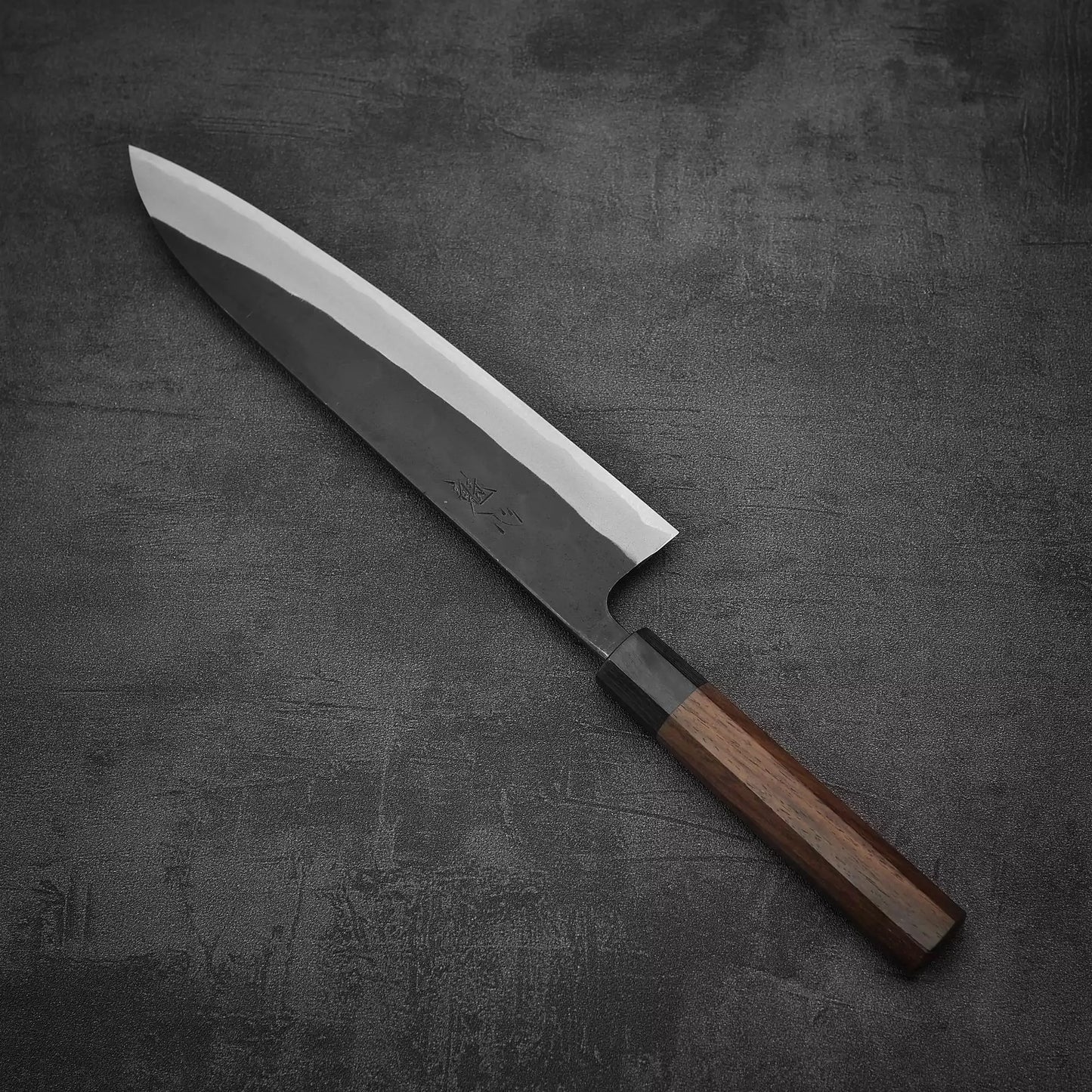 The Nao Yamamoto kurouchi shirogami#2 gyuto 240mm, featuring a sharp angular blade and dark wooden handle, is displayed on a textured dark gray surface.