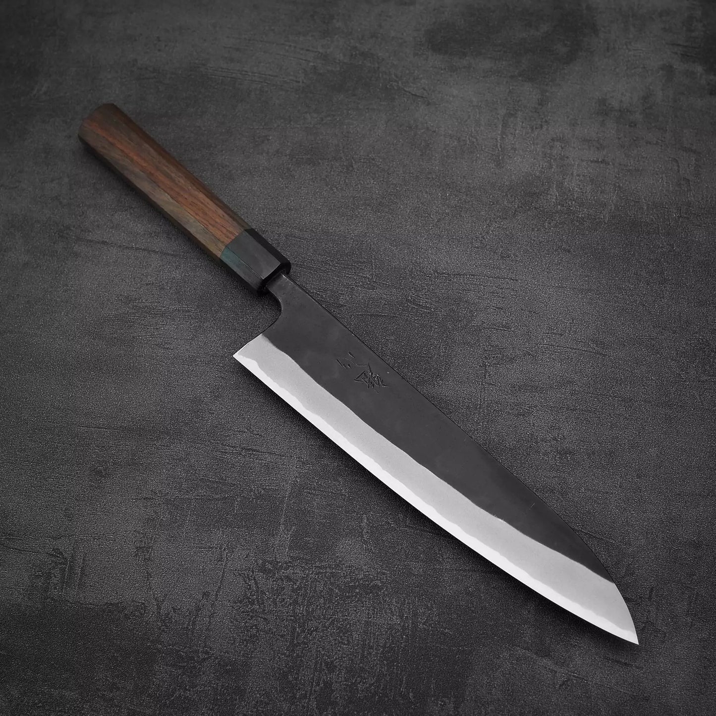 The Nao Yamamoto kurouchi shirogami#2 gyuto 240mm, a handmade Japanese knife with a dark wooden handle, rests on a textured dark gray surface.
