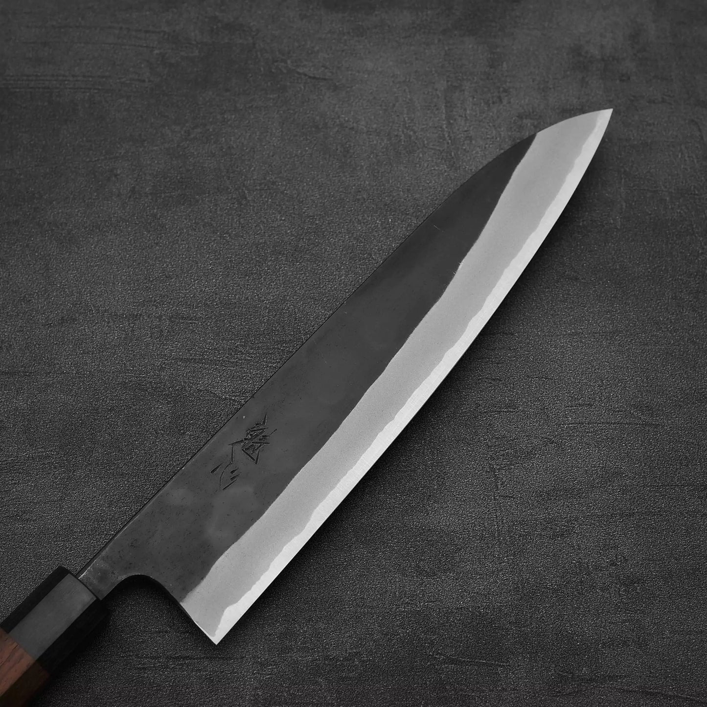 The Nao Yamamoto kurouchi shirogami#2 gyuto 240mm, a handmade Japanese knife with a dark handle and sharp polished blade, rests on a textured dark gray surface.