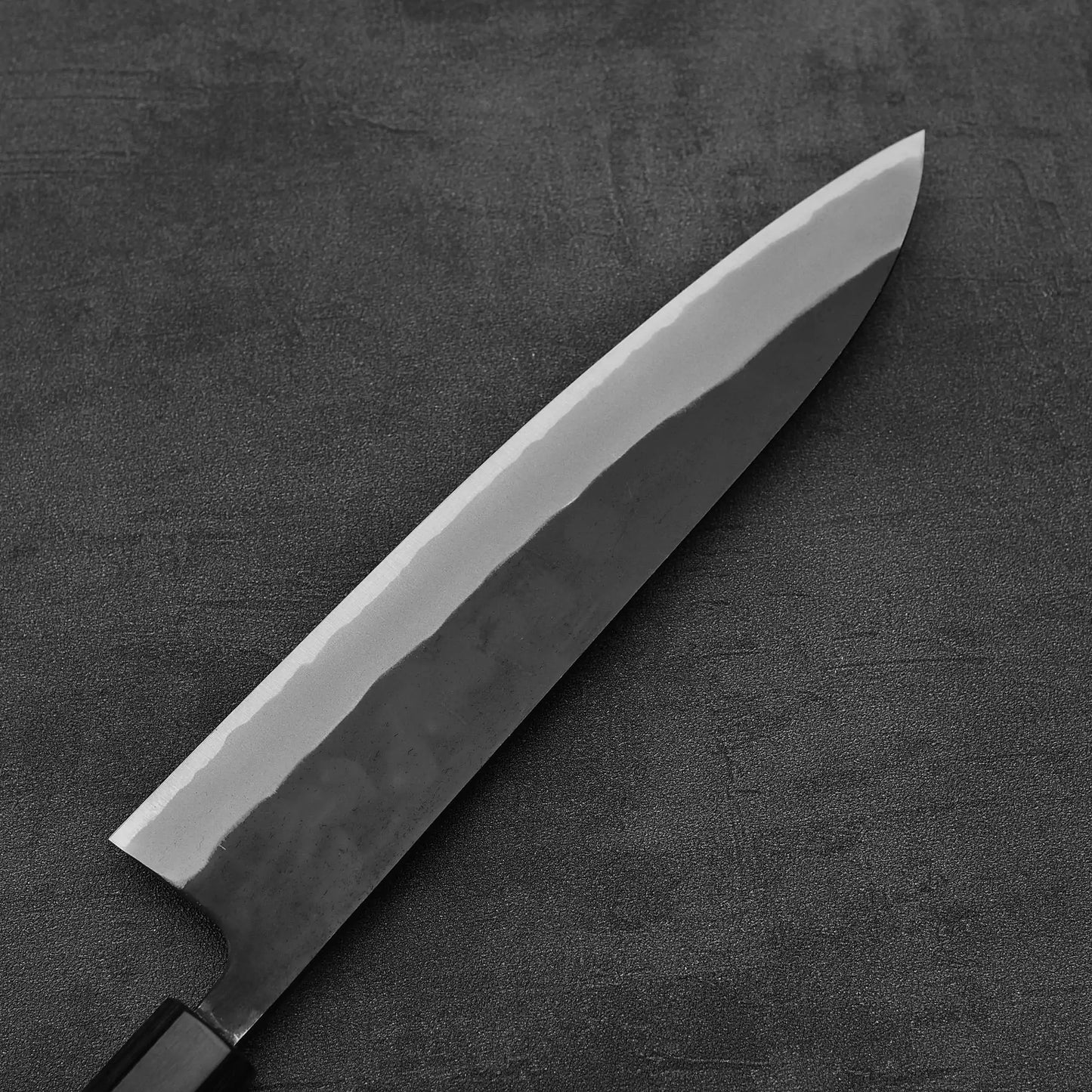 The Nao Yamamoto kurouchi shirogami#2 gyuto 240mm features a shiny, sharp blade and black handle, resting on a dark textured surface. Its polished edge reflects light beneath the kurouchi finish.