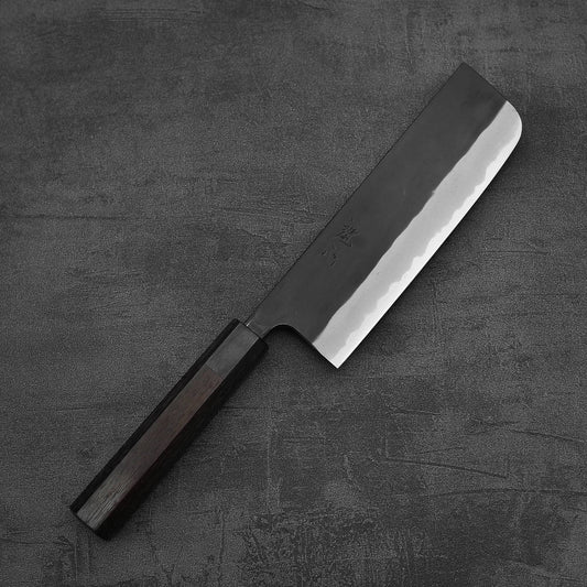 The Nao Yamamoto kurouchi shirogami#2 nakiri 165mm, a Japanese-style knife with a straight-edged blade and dark wooden handle, featuring a rustic kurouchi finish, rests on a textured dark gray surface.
