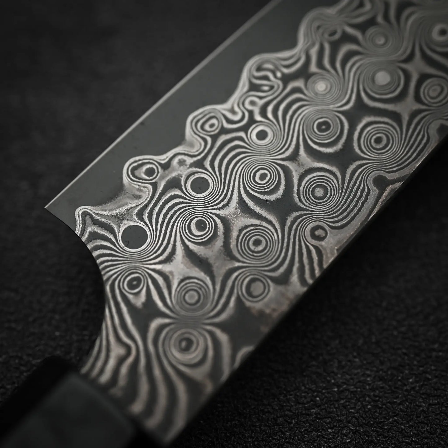 Close-up of the Nigara Hamono Nigara Anmon SG2 damascus petty knife 150mm blade, highlighting its detailed Damascus steel patterns with swirling designs and circular motifs against a dark textured background.