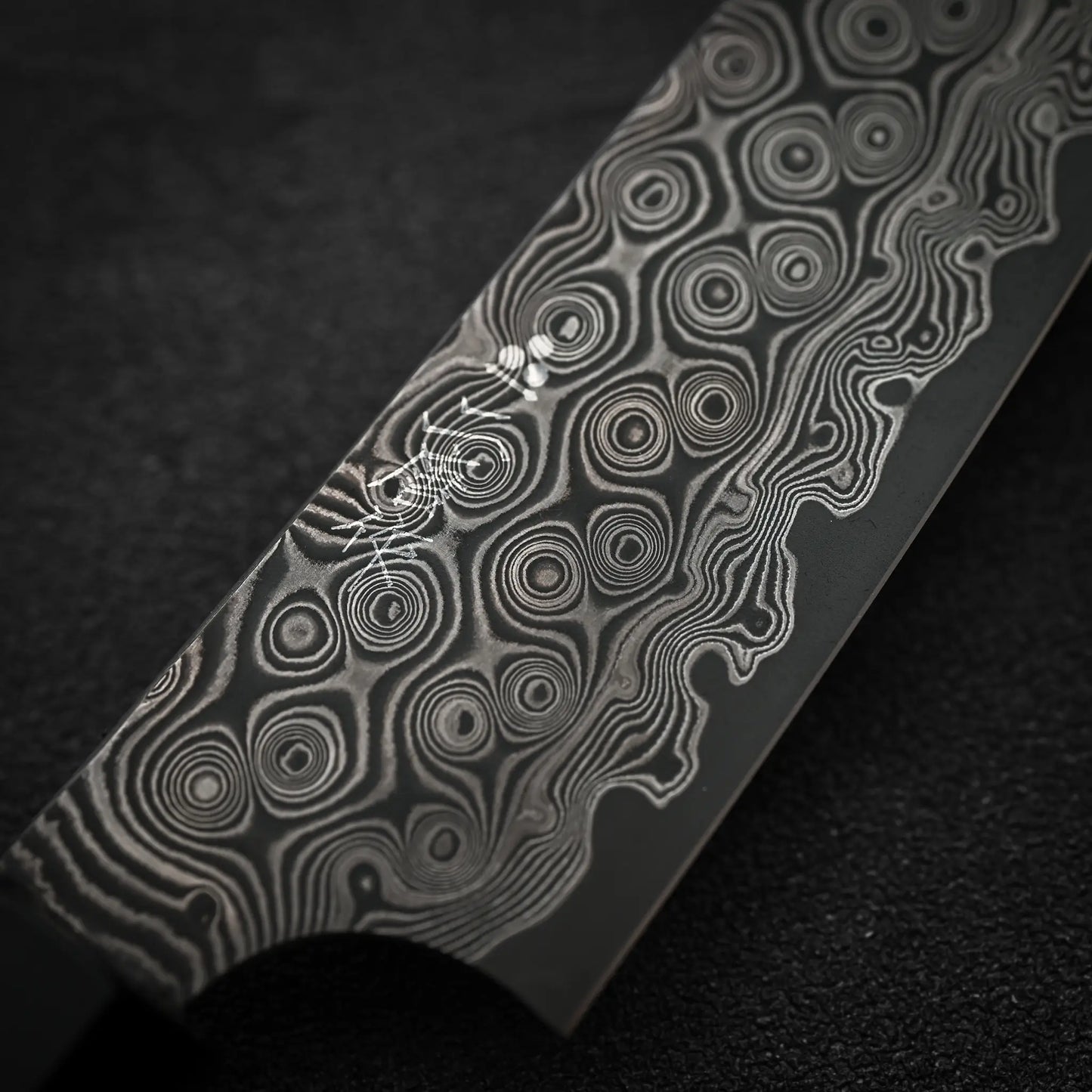 A close-up of the Nigara Hamono Nigara Anmon SG2 damascus petty knife 150mm showcases its detailed, circular wavy Damascus steel blade pattern against a black textured background.