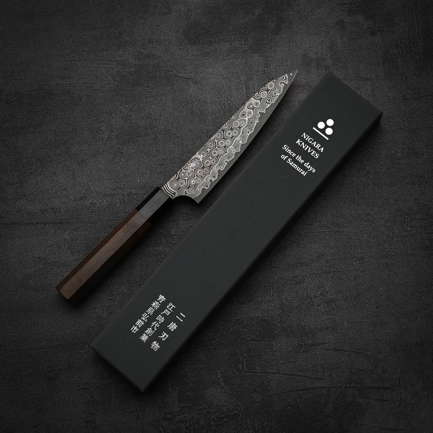 A Nigara Hamono Nigara Anmon SG2 damascus petty knife 150mm with a dark handle rests beside a black rectangular NGMA KNIVES box featuring English and Japanese text, all displayed on a dark textured surface.