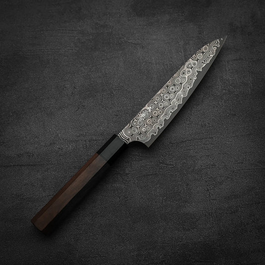The Nigara Hamono Nigara Anmon SG2 damascus petty knife 150mm, featuring a patterned Damascus steel blade and dark wooden handle, rests on a dark textured surface.